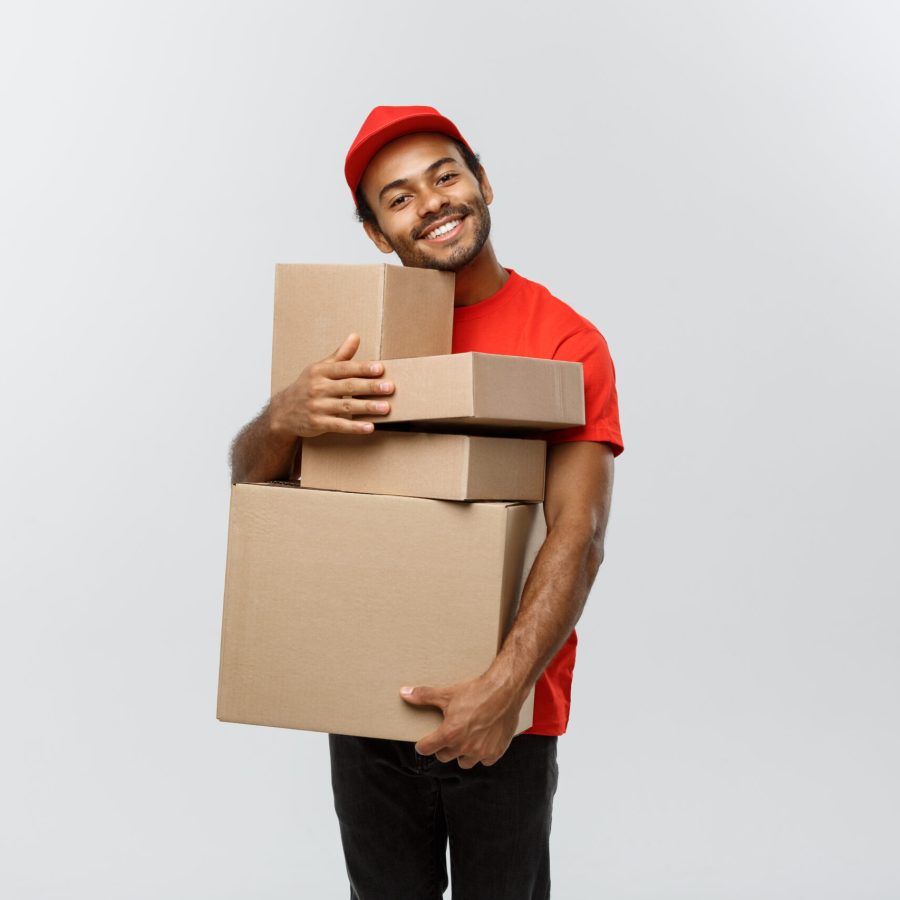 Delivery Concept – Portrait of Happy African American delivery man in red cloth holding a box package. Isolated on Grey studio Background. Copy Space. Delivery Concept - Portrait of Happy African American delivery man in red cloth holding a box package. Isolated on Grey studio Background. Copy Space.